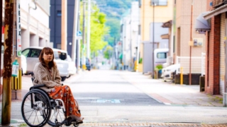 車椅子に乗った女性が街中の歩道にいる様子。背景には建物と車が見える。