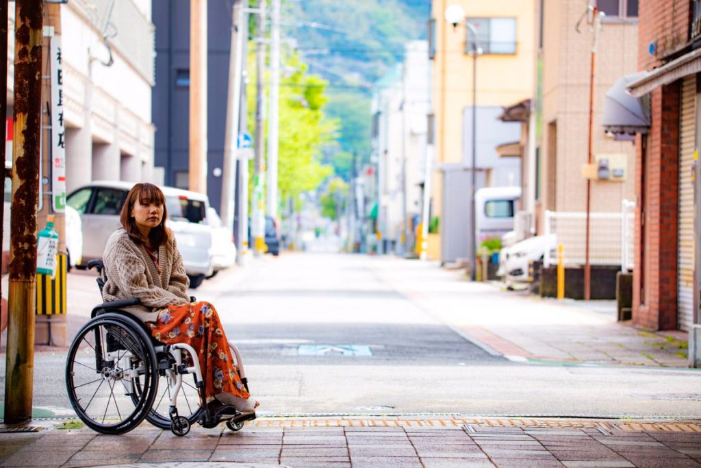 車椅子に乗った女性が街中の歩道にいる様子。背景には建物と車が見える。