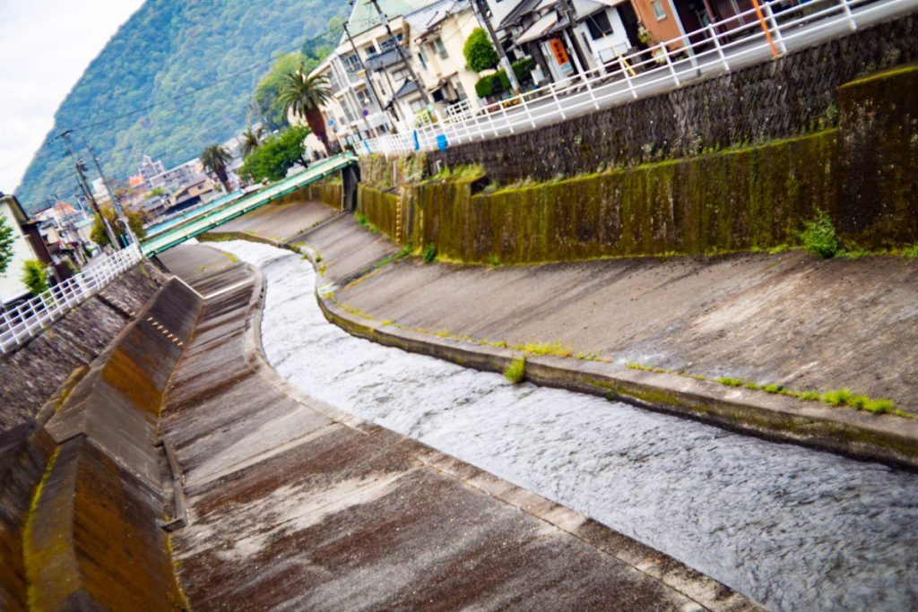 小さな川が町の中を流れており、背景には山が見える。川沿いには家や木々が並んでいる。