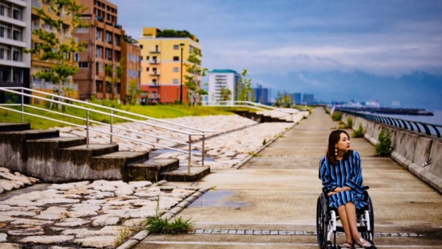 海辺を車椅子で散歩している女性。背景には建物と石畳が見える