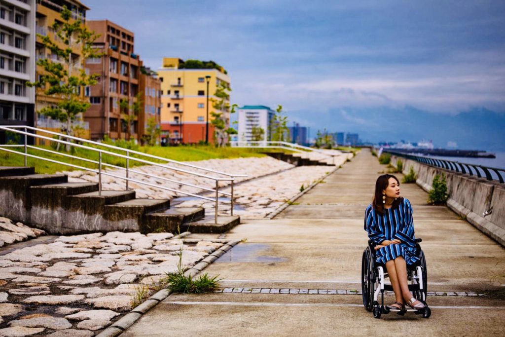 海辺を車椅子で散歩している女性。背景には建物と石畳が見える