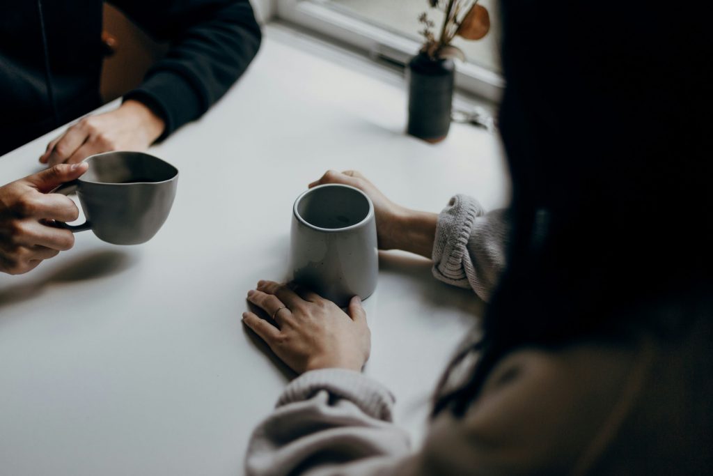 カフェのテーブルで2人が向かい合い、コーヒーカップを手にして話している様子。手元には白いカップが見える。