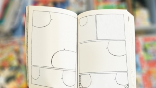 漫画のコマ割りが描かれた空白のページを持つ手。背景に他の漫画がぼんやりと写っている。