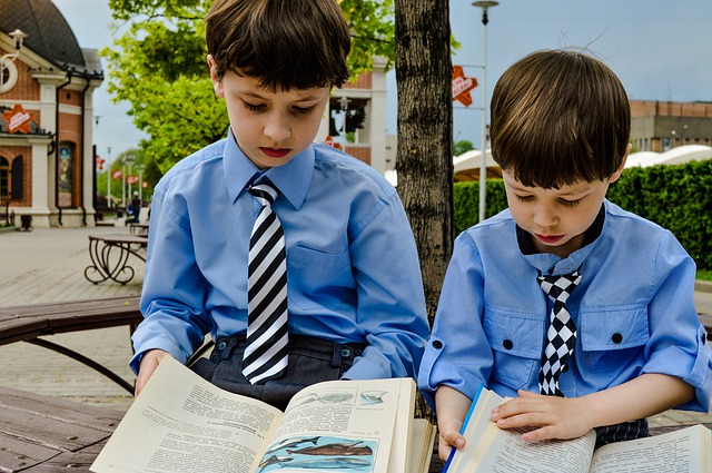 青いシャツとネクタイを着た2人の男の子が、屋外のベンチで本を読んでいる様子。