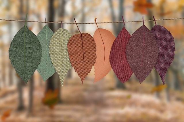 秋の背景に対して糸に吊るされた様々な色とテクスチャの布製の葉。葉は緑から赤、オレンジ、茶色までのグラデーションで並んでいる。