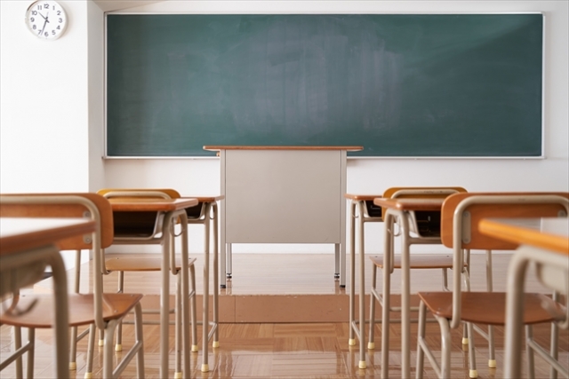 前方に黒板と教師の机があり、生徒用の机と椅子が整然と並んでいる空の教室。左上に時計が掛かっている。