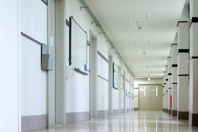 白い壁と天井、そして数多くのドアが並んでいる空の学校の廊下。奥に扉が見える。