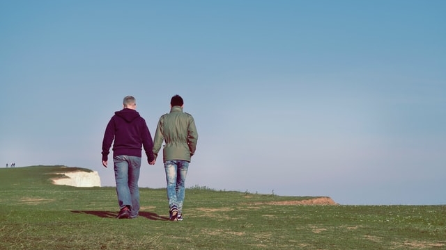 草原を歩くゲイのカップルが手をつないでいる様子。非日常と日常を象徴する場面です。