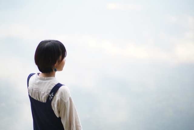 空を背景に遠くを見つめる短髪の女性の後ろ姿。