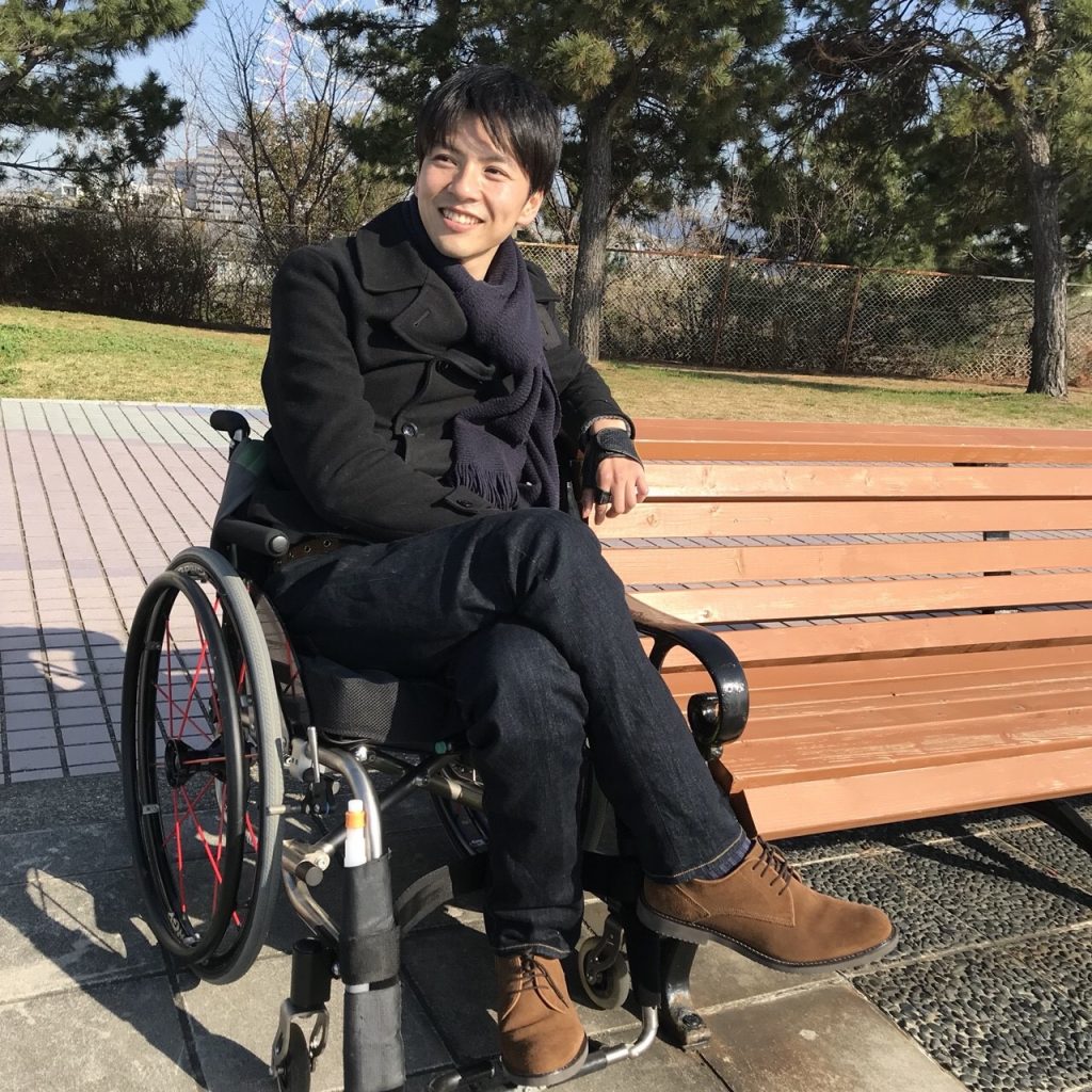 車椅子に座り、公園のベンチの横で笑顔を見せる男性。