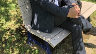スーツを着た男性が公園のベンチに座っている様子。障害を負う前の場所に戻ることの是非について考える場面を象徴しています。