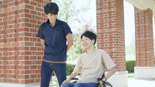 車椅子に座っている男性と立っている男性が笑顔で会話している様子。