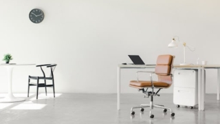 白い壁と床のシンプルなオフィススペース。茶色の椅子とデスク、ノートパソコン、ランプが配置されている。背景には時計と植物があるテーブルが見える。