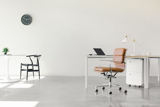 白い壁と床のシンプルなオフィススペース。茶色の椅子とデスク、ノートパソコン、ランプが配置されている。背景には時計と植物があるテーブルが見える。