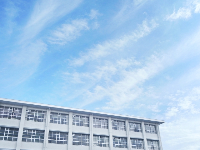 青空の下に立つ学校の建物。白い雲が広がる晴れた空が背景にあり、建物の窓が整然と並んでいる。