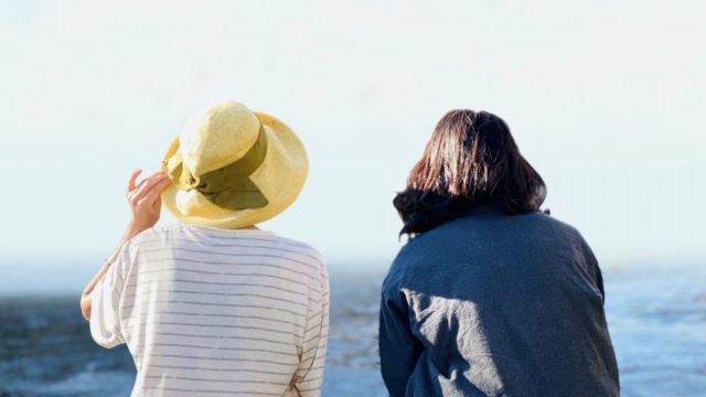 海を背景に並んで座る、麦わら帽子をかぶった女性と黒髪の女性の後ろ姿。