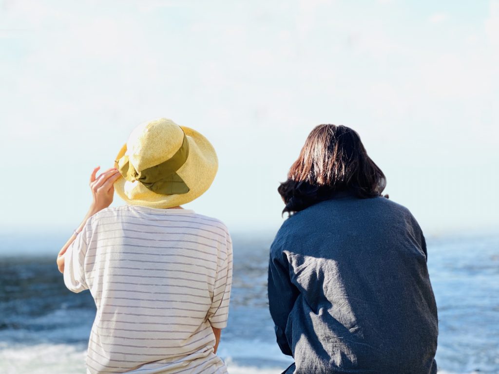 海を背景に並んで座る、麦わら帽子をかぶった女性と黒髪の女性の後ろ姿。
