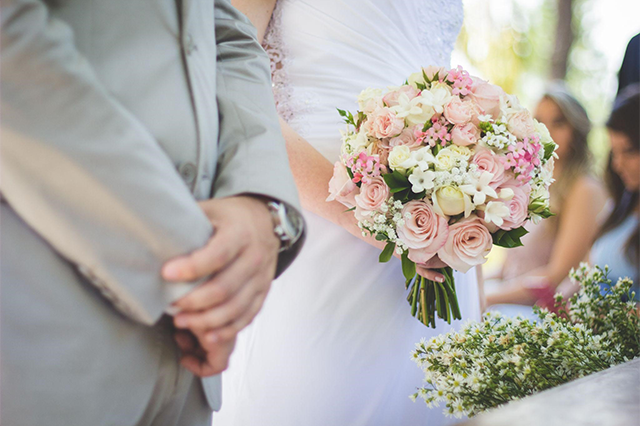 結婚式の様子。花嫁がピンクと白の花束を持ち、花婿がその隣に立っている。