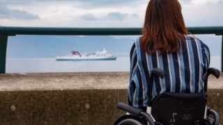車椅子に座って海を見つめる女性の後ろ姿。遠くには船が見える。