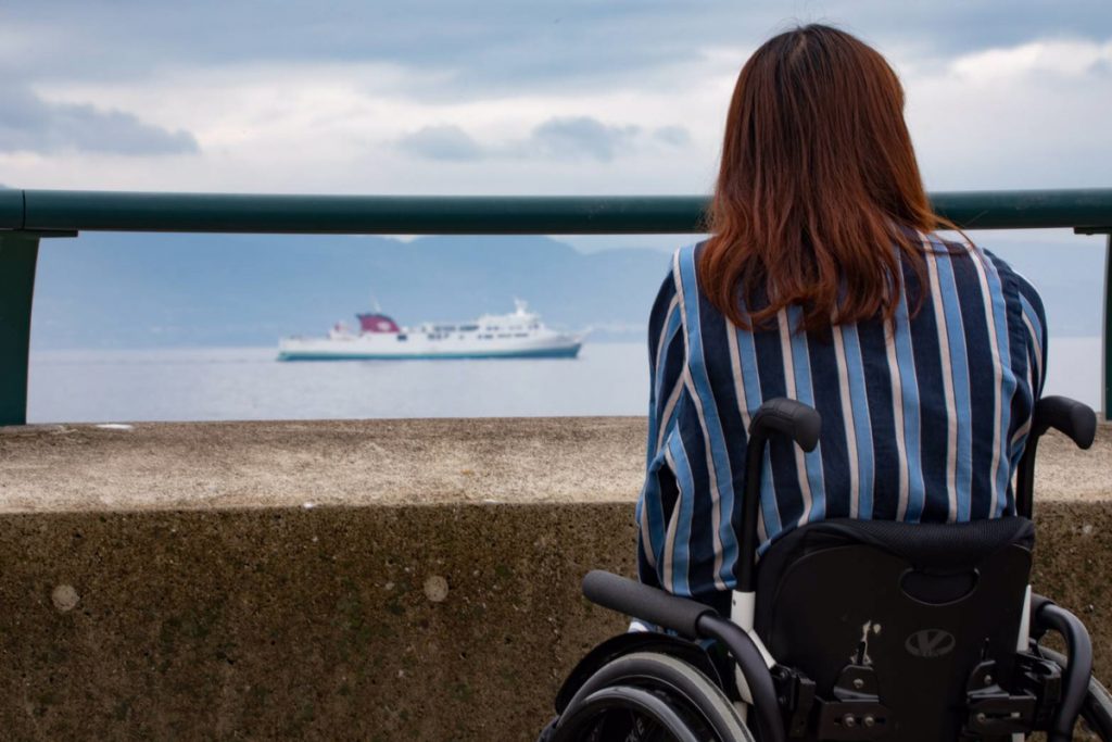 車椅子に座って海を見つめる女性の後ろ姿。遠くには船が見える。