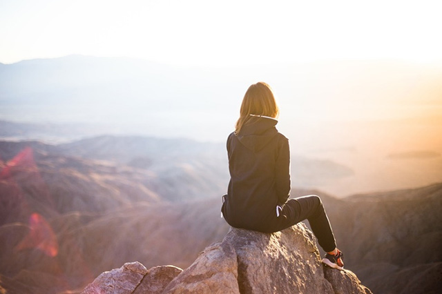 夕日を背景に山の頂上で座って景色を眺める女性。