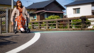 車椅子に座って道を進む女性、背景には住宅街が広がっている。