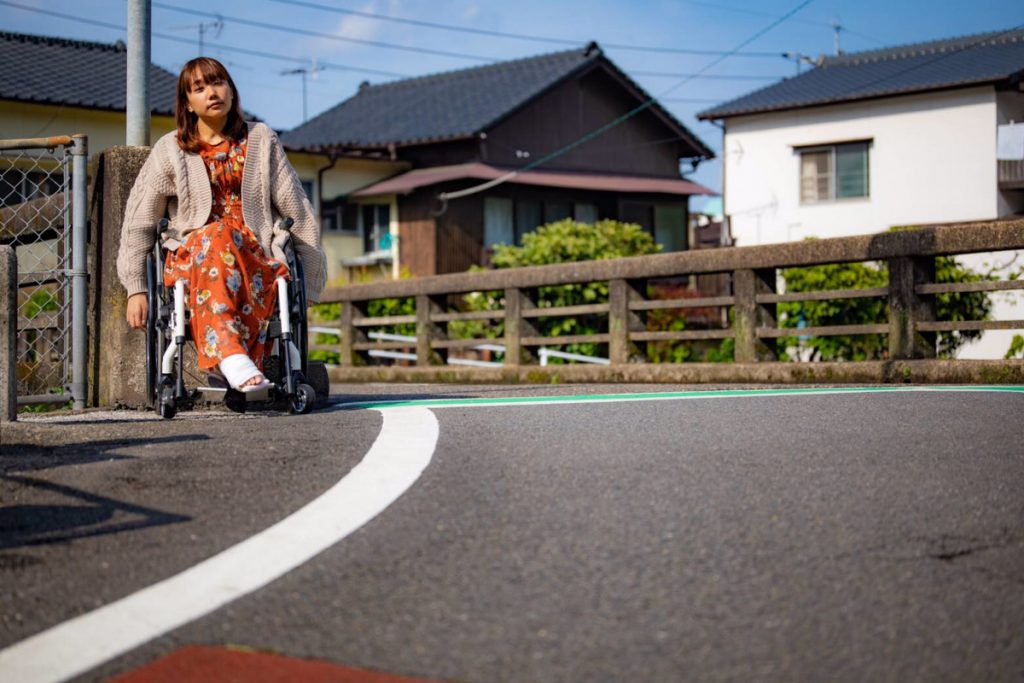 車椅子に座って道を進む女性、背景には住宅街が広がっている。