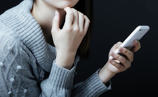 グレーのセーターを着た若い女性がスマートフォンを持ち、考え込んでいる様子。21歳で生活保護を受給する20代の難病患者が生活困窮時に支援を求める場面を示す画像。